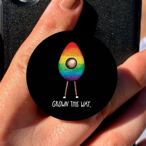 Pode incluir: Uma capa de telefone preta com um suporte de telefone de abacate arco-íris. O abacate está usando um arco-íris e tem o texto "Grown This Way."