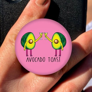 Pode incluir: Um pop socket rosa para telefone com um gráfico de duas metades de abacate segurando copos de líquido verde. O texto "Avocado Toast" está impresso abaixo dos abacates.