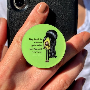 Pode incluir: Um agarre de telefone verde com uma ilustração de desenho animado de uma mulher com cabelo preto e um vestido amarelo. O texto no agarre diz "Eles tentaram me fazer ir para reabilitação, mas Hass disse não, não, não."