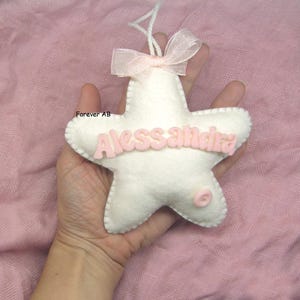 Può includere: Una stella di feltro bianco con il nome "Alessandra" in lettere di feltro rosa. La stella ha un bottone rosa e un nastro rosa.