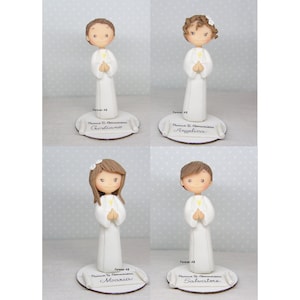 Può includere: Quattro piccole figurine di bambini vestiti con abiti bianchi da comunione. Le figurine sono in piedi su piccole basi di legno con rotoli che hanno i nomi "Cristiano", "Angelica", "Mooxia" e "Salvatore" scritti su di essi.
