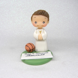 Può includere: Una statuina bianca di un bambino che indossa una tunica bianca e tiene le mani giunte in preghiera. Ha i capelli castani e un pallone da basket marrone ai suoi piedi. La base della statuina è verde e ha il testo "Prima S. Comunione Leonardo" scritto su di essa.