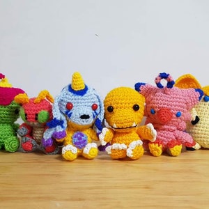 May include: A collection of eight crocheted stuffed toys, each featuring a different character design. The toys are made in a variety of colors, including white, green, pink, yellow, and blue.