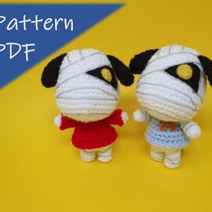 May include: Two crocheted mummy dog plushies, one wearing a red dress and the other wearing a blue dress. The plushies have black ears and yellow eyes. The background is yellow. Pattern PDF.
