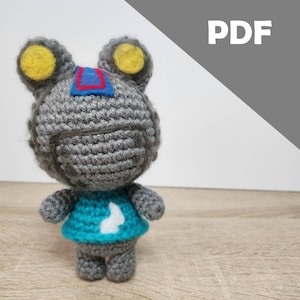 May include: A gray crocheted frog amigurumi toy with yellow eyes and a blue and red stripe on its head. The frog is wearing a teal crocheted shirt with a white design.