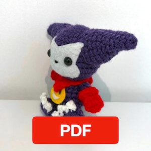 May include: A handcrafted crochet doll featuring a dark purple head with pointed ears, a white face, and black button eyes. The body is dark purple with a red collar, red sleeves, white accents on the feet, and a yellow circular emblem on the chest.