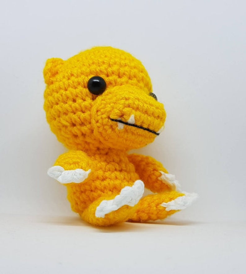 K&ouml;nnte beinhalten: Ein gelbes H&auml;kel-Dinosaurierspielzeug mit wei&szlig;en Krallen und schwarzen Augen.