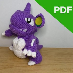 Puede incluir: Un dinosaurio morado tejido a mano con detalles blancos, incluyendo un vientre, garras y dientes blancos. El dinosaurio tiene un cuerno, detalles de orejas amarillas y verdes y una pequeña cola. La palabra "PDF" está en un cuadro verde en la esquina superior derecha.
