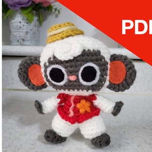 Pode incluir: Um brinquedo de pelúcia de crochê de uma ovelha usando um chapéu amarelo e uma camisa vermelha com um desenho de flor. A ovelha tem olhos pretos e um nariz cinza.