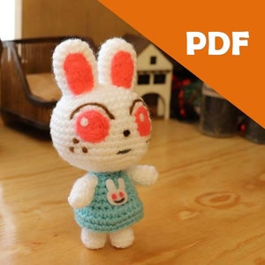 May include: White crocheted bunny rabbit plush toy with pink ears and red eyes wearing a light blue dress with a white bunny graphic.