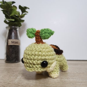 May include: A crocheted green turtle with a brown shell and a green leafy sprout on its back. The turtle has black eyes and a white belly.
