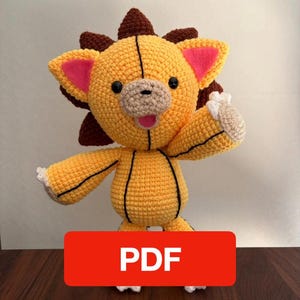 May include: A handmade crochet lion plush toy in yellow, brown, and pink. The lion has a smiling face, a brown mane, and is holding up one paw. The word "PDF" is displayed on a red rectangle at the bottom.