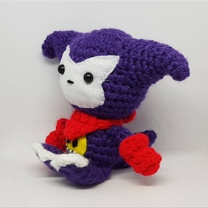 May include: A purple crocheted plush toy featuring a white felt face mask, black bead eyes, and a red scarf. The figure has red hands and feet, with a yellow circular emblem on the chest displaying a white skull graphic and white yarn accents.