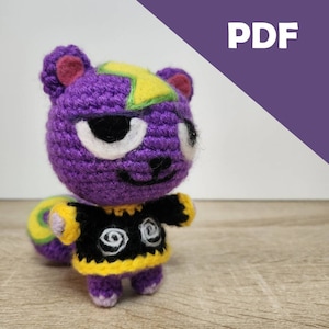 May include: Crocheted purple animal plush toy with yellow and green accents. The toy is wearing a black and yellow sweater with a white swirl design.