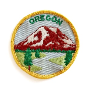 May include: Embroidered patch featuring a mountain scene with the word "OREGON" in green. The mountain is red and white, with green trees and a light blue background. The patch has a yellow border.