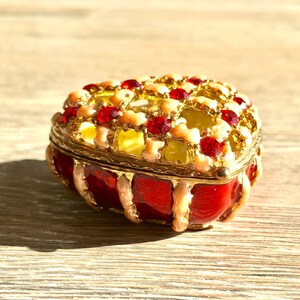 May include: A red heart-shaped trinket box with a gold lattice design and red gem accents. The box is closed and sitting on a wooden surface.