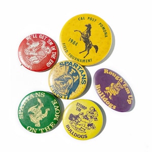May include: Six vintage button pins with various designs and colors. The pins feature images of a horse, a mascot, and a bulldog. The text on the pins includes "Cal Poly Pomona", "1984", "Field Tournament", "Spartans", "Get 'Em In The End", "Matadors", "Rough 'Em Up", "Bulldogs", "First", "Stomp 'Em", "On The Move".