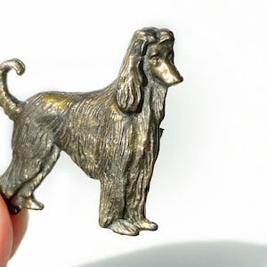 May include: A small, bronze-colored figurine of an Afghan Hound dog. The dog is standing with its tail curled up and its head turned to the side. The figurine is detailed with a textured surface to represent the dog's fur.