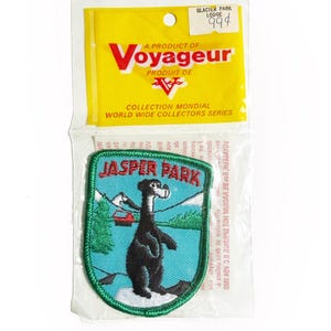 May include: A vintage Jasper Park patch featuring a black bear, a red ski lift, and the text "JASPER PARK" on a teal background. The patch is in a clear plastic package with a yellow header that reads "Voyageur".