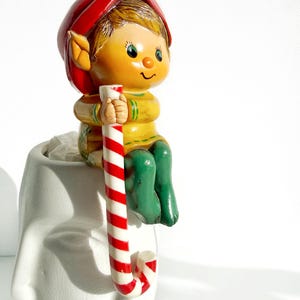 May include: A ceramic Christmas elf figurine with a red hat, yellow shirt, and green pants, holding a red and white striped candy cane. The elf is perched on a white face-shaped planter.