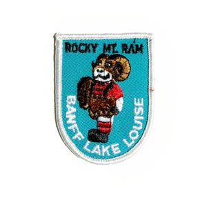May include: A patch featuring a cartoon ram in a red shirt and black boots. The patch has a teal background with the words "ROCKY MT. RAM", "BANFF", and "LAKE LOUISE" in white lettering. The ram has brown fur and large horns.