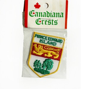 May include: A patch from the "Canadian Crests" collection, featuring the Prince Edward Island coat of arms. The patch is in a clear plastic sleeve, with a red and white header. The crest includes a lion, trees, and text.