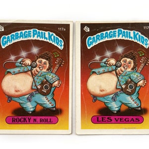 May include: Two Garbage Pail Kids trading cards featuring cartoon characters with the text "Garbage Pail Kids" and the numbers "117a" and "117b". The first card features a character named "Rocky N. Roll" and the second card features a character named "Les Vegas".
