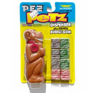 May include: A brown plastic "Grinz" the monkey Pez dispenser with a red button on its chest. The dispenser comes with a pack of Amurol bubble gum in assorted flavors: fruit, grape, and watermelon.