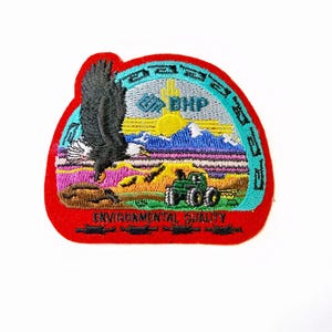 May include: Embroidered patch with a red border featuring a landscape scene. The design includes a soaring eagle, mountains, a tractor, and the text "ENVIRONMENTAL QUALITY" and "BHP". The patch has a colorful design with blue, green, yellow, and purple hues.