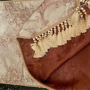 May include: A beige and brown table runner with a gold tassel trim. The runner has a floral pattern and is reversible.