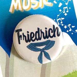 May include: A white button with the text "Friedrich" in black lettering and a blue neckerchief design. The button is on a card with the text "The Sound of Music" in yellow and blue lettering.