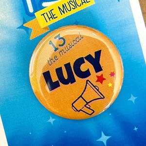 May include: A blue and white card with a large white number 13 and the text "13 The Musical". A yellow button with the text "13 the musical Lucy" and a megaphone graphic is attached to the card.