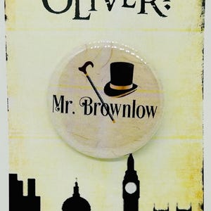 May include: A framed print featuring the word "OLIVER!" in bold black lettering. A circular badge with a top hat and cane design is attached, with "Mr. Brownlow" below. A black silhouette of a city skyline is at the base of the print.