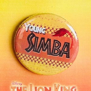 Puede incluir: Un botón amarillo y naranja con el texto "Young Simba" y un diseño de llama roja. El botón está en una tarjeta con el texto "Disney's The Lion King Jr."
