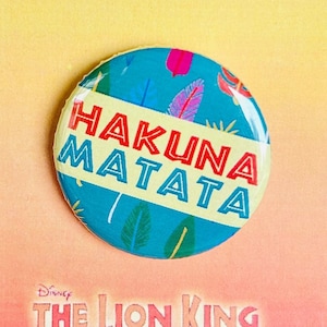 Puede incluir: Un botón redondo con fondo azul y hojas y plumas de colores. El botón dice "Hakuna Matata" en letras amarillas y rojas. El botón está en una tarjeta con las palabras "Disney El Rey León Jr." en letras rojas y amarillas.