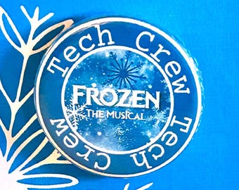 May include: A blue card with the words "FROZEN The Musical" in white script. A round pin with "Tech Crew" and "FROZEN The Musical" is affixed to the card. A white snowflake design is in the lower left corner.