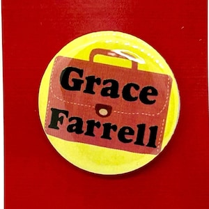 May include: A red and white card with the text "Annie the musical." A yellow circle with a red briefcase graphic and the name "Grace Farrell" is centered on the card. The card is on a black surface.