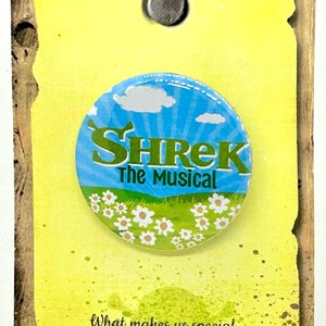 May include: A yellow card with a green, blue and white button featuring the text "Shrek The Musical". The card has a quote from the musical: "What makes us special, makes us strong. -Shrek"