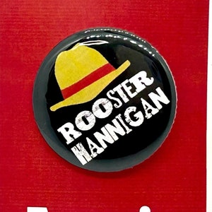 May include: A black and white button with a yellow and red hat and the text "Rooster Hannigan" on a red background with the text "Annie the musical" below the button.