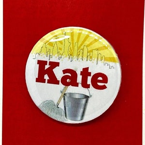 Könnte beinhalten: Eine rote und weiße Karte mit einem gelben Kreis in der Mitte. Der Kreis hat eine weiße Silhouette einer Stadtkulisse und das Wort "Kate" in Schwarz. Der Kreis hat auch einen Besen und einen Eimer. Die Karte trägt die Worte "Annie das Musical" in weißen Buchstaben.