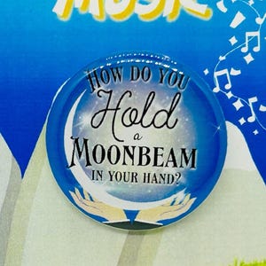 May include: A rectangular card with a blue sky background and the words "THE SOUND OF MUSIC" in yellow. A round button with the text "HOW DO YOU HOLD A MOONBEAM IN YOUR HAND?" is attached to the card. White mountains and green grass are also visible.