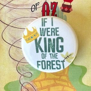 Peut inclure: Un bouton blanc avec un motif vert et bleu qui dit "Si j'étais roi de la forêt". Une couronne dorée est au-dessus du texte. Le bouton est sur une route de briques jaunes avec une montgolfière en arrière-plan.