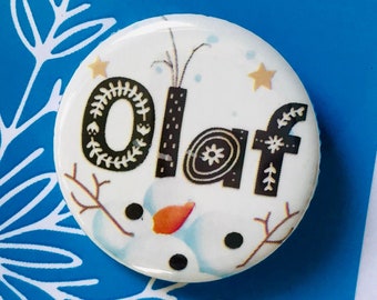 May include: A white button with a black and white illustration of Olaf from Frozen The Musical. The button has the word "Olaf" written in black with white snowflakes and stars.