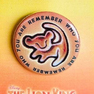 Puede incluir: Un pin redondo con un cachorro de león de dibujos animados dentro de un círculo. El texto alrededor del cachorro de león dice "Remember who you are who you are remember". El pin está en una tarjeta con el texto "Disney The Lion King Jr."
