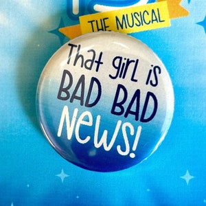 May include: A blue and white button with the text "That girl is BAD BAD NEWS!" on a white background with a blue and yellow border. The button is on a card with the number "13" and the text "THE MUSICAL" in yellow.