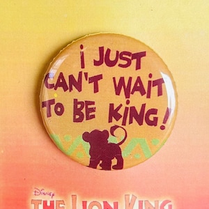 以下が含まれることがあります： 「I Just Can't Wait To Be King!」という文字と、漫画のライオンの子が描かれた黄色とオレンジのボタン。ボタンは、「Disney The Lion King Jr.」という文字が書かれた黄色とオレンジのカードの上に置かれています。