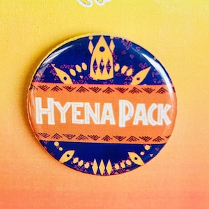 Puede incluir: Un botón redondo con un diseño estampado azul y naranja. El botón dice "HYENA PACK" en texto blanco. El botón está sobre un fondo amarillo y naranja con el texto "THE LION KING KIDS" en la parte superior.