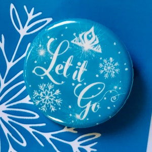 May include: A blue button with a white snowflake design and the text "Let it Go" in white cursive lettering. The button also features a white crown and sparkles. The button is on a blue background with a white snowflake design.
