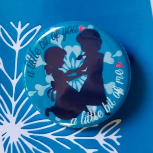 May include: A blue button with a silhouette of two people holding hands. The text on the button reads "A little bit of you, a little bit of me".