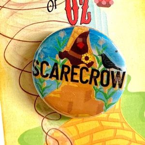 Peut inclure: Un bouton jaune, bleu et vert avec une image de dessin animé de l'Épouvantail du Magicien d'Oz. Le bouton indique "Scarecrow" en texte noir. Le bouton est sur une carte avec le texte "We're off to see the Wizard... The Wonderful... Wizard of Oz".